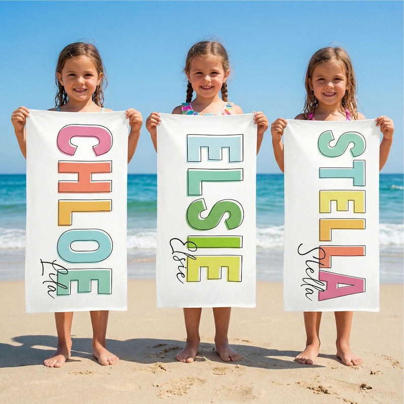 May include: Three white beach towels with colorful names: "CHLOE", "ELSIE", and "STELLA". Each name is in a different color and font, with a smaller name below. The towels are held by three young girls on a sandy beach with the ocean in the background.
