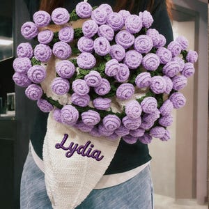 May include: A handmade crochet bouquet featuring numerous lavender-colored roses. The roses are arranged with green crocheted leaves and a white crocheted cone-shaped holder. The name "Lydia" is embroidered in purple script on the holder.