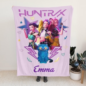 May include: A light purple blanket with the text "HUNTRAX" and the name "Emma". The design includes a graphic of three animated women with microphones and a blue cat. Decorative stars and lightning bolts are also present.