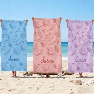 May include: Three beach towels in blue, coral, and lavender, each featuring a seashell pattern and the name "Susan" in script. The towels are held up on a sandy beach with the ocean in the background.