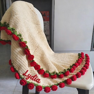 May include: Cream-colored crocheted blanket with a red rose border and the name "Lydia" embroidered in red. The blanket is draped over a white chair, showcasing the intricate floral design and handmade quality.