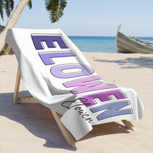 May include: White beach towel draped over a wooden beach chair. The towel features the word "FLOWEN" in purple and lavender block letters, with "Flowen" in cursive below. The scene is set on a sandy beach with a boat and palm tree.