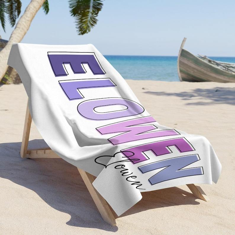 May include: White beach towel draped over a wooden beach chair. The towel features the word "FLOWEN" in purple and lavender block letters, with "Flowen" in cursive below. The scene is set on a sandy beach with a boat and palm tree.