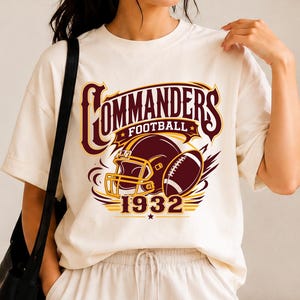 May include: An off-white short-sleeve t-shirt featuring a graphic print in burgundy and yellow. The design includes the text COMMANDERS FOOTBALL, a football helmet, a football, and the year 1932 in a bold, stylized font across the chest.