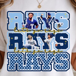 Può includere: T-shirt bianca con un design blu e azzurro che presenta la parola "RAYS" in formato impilato. Il design include immagini di giocatori di baseball e il testo "let's go rays".