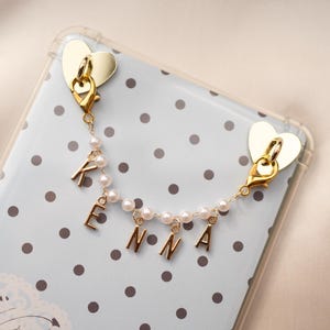 May include: A phone case with a light blue polka dot design and a gold heart-shaped charm. A pearl and gold chain spells out the name "KENNA". The phone case is clear.