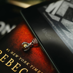 May include: A close-up of a book and an e-reader. The book has a red cover with gold lettering that reads "REBECCA" and "#1 NEW YORK TIMES". A silver bookmark with a claw design and a gold bead is on the book. The e-reader is black and has the word "kindle" on it.