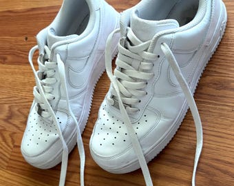 May include: A pair of white leather low-top sneakers with white laces. The sneakers are on a wooden surface.