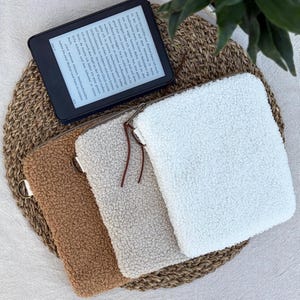 Könnte beinhalten: Drei rechteckige Taschen aus weichem, strukturiertem Sherpa-Fleece in Weiß, Beige und Braun. Jede Tasche verfügt über einen Metallreißverschluss mit einem dünnen braunen Lederanhänger. Ein schwarzer E-Reader mit deutschem Text liegt darüber.