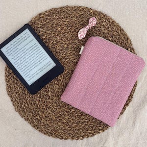 Könnte beinhalten: Ein schwarzer E-Reader mit deutschem Text neben einer rechteckigen, gesteppten Stoffhülle in Altrosa mit kleinen weißen Punkten und einer dekorativen Schleife aus rosa-weißem Blumenstoff an einem Metallring am oberen Rand.