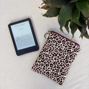 Könnte beinhalten: Ein schwarzer E-Reader, der den Text Ashlyn in Cornwall mit einem deutschen Textabschnitt darunter anzeigt, neben einer rechteckigen Stofftasche mit braunem, schwarzem und cremefarbenem Leopardenmuster und einem dunkelroten Reißverschluss.