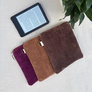 Könnte beinhalten: Drei rechteckige Etuis aus Kord in Dunkelbraun, Hellbraun und Dunkellila, jeweils mit einem seitlichen Reißverschluss und einem Metallring als Zipper. Ein schwarzer E-Reader mit deutschem Text auf dem Display liegt oberhalb der Etuis.