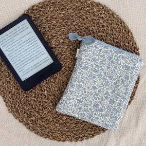 Könnte beinhalten: Ein schwarzer E-Reader mit deutschem Text neben einer rechteckigen Stofftasche mit hellblau-weißem Blumenmuster. Die Tasche hat einen passenden Reißverschlussanhänger mit Punkten und einen kleinen, seitlich angebrachten Metall-D-Ring.