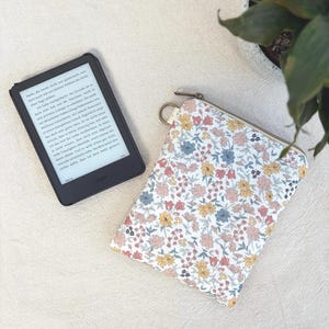 Könnte beinhalten: Ein schwarzer E-Reader mit einem Bildschirm, der deutschen Text anzeigt, und eine rechteckige Stofftasche mit Blumenmuster in Gelb-, Rosa-, Blau- und Grüntönen, ausgestattet mit einem Reißverschluss und einem kleinen goldenen Metallring an der Seite.