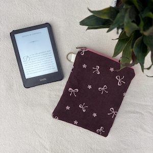 Könnte beinhalten: Ein schwarzer Kindle E-Reader mit dem Text Ashlyn in Cornwall neben einer rechteckigen, dunkelvioletten Stofftasche mit einem wiederkehrenden Muster aus kleinen hellrosa Schleifen und Blumen, ausgestattet mit einem Reißverschluss.
