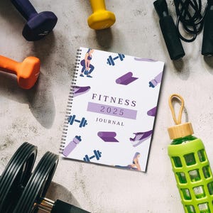 May include: A white fitness journal with a purple and white design featuring a woman exercising. The journal has a spiral binding and the text "Fitness 2025 Journal". The journal is surrounded by various fitness equipment, including dumbbells, a jump rope, and a water bottle.