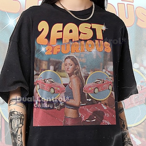 May include: Black unisex t-shirt featuring the text "2 Fast 2 Furious" in orange and yellow. The graphic includes an image of a woman and two red convertibles. The shirt also has the text "Dual Control* On Etsy".