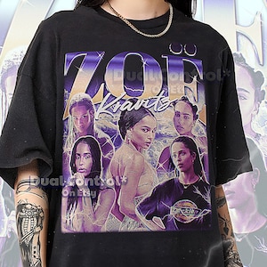 Op de afbeelding: Zwart unisex T-shirt met een grafische afbeelding van de naam "Zoe Kravitz" in paars en goud, met afbeeldingen van Zoe Kravitz. Het shirt heeft een vintage uitstraling. De tekst "Dual Control" is ook zichtbaar.