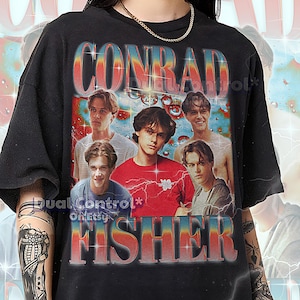 Peut inclure: T-shirt unisexe noir avec un design rétro affichant le nom "CONRAD FISHER" dans une police vintage colorée. Le t-shirt présente un collage d'images de jeunes hommes. Le texte en bas fournit des informations sur les tailles.