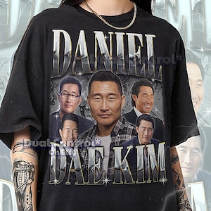 May include: Black unisex t-shirt featuring a graphic of Daniel Dae Kim with the name "DANIEL DAE KIM" in silver lettering. The shirt includes a collage of images of the actor. The text at the bottom reads "THE FIRST MODEL IS WEARING A 2XL SHIRT..."