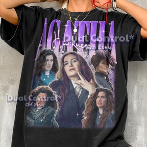 Op de afbeelding: Zwart T-shirt met een afbeelding van een vrouw met lang bruin haar, met de tekst "AGATHA Harkness" in paars en wit. De afbeelding bevat meerdere afbeeldingen van de vrouw. Het shirt is donker van kleur.