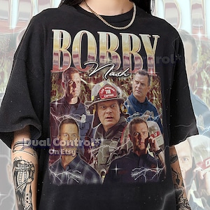 以下が含まれることがあります： 消防士のヴィンテージスタイルのグラフィックが特徴の黒いTシャツで、「BOBBY Nash」の文字が入っています。デザインには消防士の画像と「Los Angeles 118 Captain」の文字が含まれています。