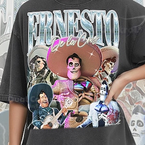 May include: Dark gray t-shirt featuring a graphic of Ernesto de la Cruz from the movie Coco. The design includes multiple characters in vibrant colors, with the name "Ernesto de la Cruz" in a stylized font. The shirt is labeled "Comfort Color Pepper".