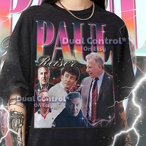 Op de afbeelding: Zwart unisex T-shirt met een afbeelding van Paul Reiser in retrostijl. Het shirt heeft de tekst "PAUL REISER" in een verlooplettertype en de Etsy shopnaam "Dual Control On Etsy".