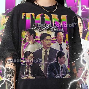 May include: Black unisex t-shirt featuring a graphic of Tom Pelphrey with the text "Tom Pelphrey" in a retro style. The shirt has a crew neck and short sleeves. The text "Dual Control Etsy" is also visible.