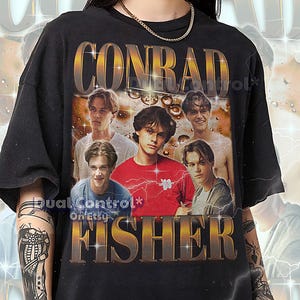 Peut inclure: T-shirt noir unisexe avec l'inscription dorée "CONRAD FISHER" et des images de jeunes hommes. Le t-shirt porte également la mention "Dual Control* On Etsy". Ce t-shirt surdimensionné taille normalement.