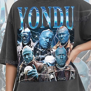 May include: A charcoal gray short-sleeve t-shirt featuring a graphic print of the character Yondu in multiple poses. The text YONDU appears in large, metallic blue letters at the top. The design includes lightning bolts and a dark, starry background.