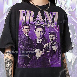 May include: Black t-shirt featuring a purple graphic of Franz Kafka and several portraits. The text "FRANZ KAFKA" is displayed in a purple, stylized font. The shirt has a crew neck and short sleeves. The text at the bottom of the shirt says: THE FIRST MODEL IS WEARING A 2XL SHIRT.