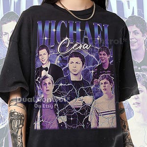 May include: Black unisex t-shirt featuring a graphic of Michael Cera with the text "MICHAEL Cera" in a vintage style. The design includes multiple images of the actor with lightning bolt accents. The shirt has a crew neck and short sleeves.