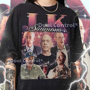 May include: Black graphic t-shirt featuring multiple images of J.K. Simmons with the text "Dual Control Simmons". The shirt includes text at the bottom. The model is wearing a 2XL shirt. This unisex shirt fits true to size.
