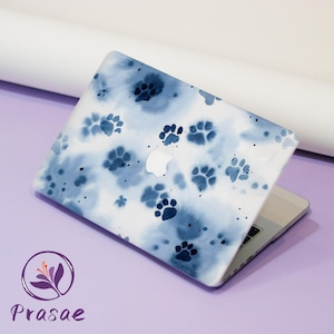 May include: A laptop with a white and blue paw print design. The laptop is open, displaying the Apple logo. The design features watercolor-style paw prints on a white background. The laptop is on a purple surface.