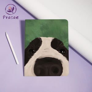 May include: A rectangular notebook featuring a close-up illustration of a dog's face with a large black nose, white muzzle, and black patches around the eyes against a green background. A white stylus is positioned to the left of the notebook.