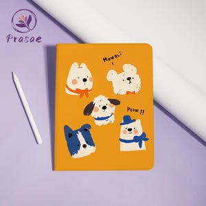 Könnte beinhalten: Ein orangefarbenes Notizbuch mit Cartoon-Hunde-Illustrationen, einige mit Schleifen und Hüten. Auf dem Notizbuch steht der Text "Mowoli" und "Peow !!". Ein weißer Stift befindet sich links vom Notizbuch.
