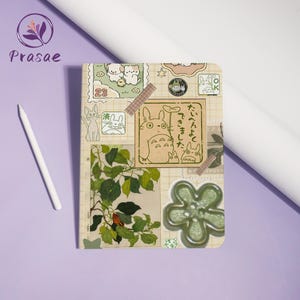 May include: A beige notebook with a collage of stickers featuring cartoon characters, plants, and Japanese text. A white stylus rests on the left. The notebook has a grid pattern and is decorated with washi tape.