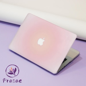 Puede incluir: Un ordenador portátil rosa con el logotipo de Apple en la tapa. El portátil está abierto, mostrando el teclado y la pantalla. El fondo es de color lila claro. El logotipo de Prasae está en la esquina inferior izquierda.