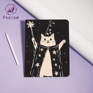 May include: Black notebook featuring a white cat dressed as a wizard, holding a star-topped wand. The cat wears a pointed hat and cape adorned with stars. A white stylus is positioned to the left. The notebook has rounded corners.