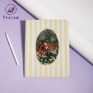 May include: A rectangular notebook with a light yellow and white vertical striped cover. The center features an oval illustration of a brown foal resting in a field of white daisies. A white stylus is positioned to the left of the notebook.