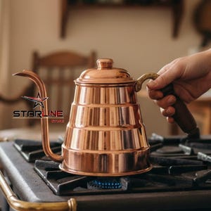 May include: A copper-colored kettle with a long, curved spout and a wooden handle sits on a black stovetop. The kettle has a tiered design and is positioned over a lit gas burner. The handle is held by a person.