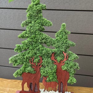 Könnte beinhalten: Eine dekorative Weihnachtsdekoration aus Holz mit zwei grünen, moosbedeckten Tannen und drei braunen Hirschsilhouetten. Die Szene steht auf einer roten Holzbasis mit weißen Akzenten und schafft eine festliche Weihnachtsdekoration.