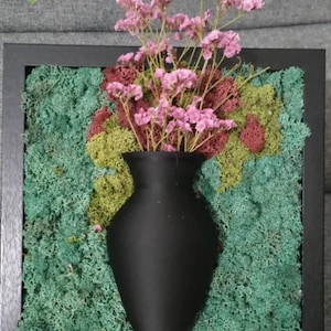 May include: A framed wall art piece featuring a black vase filled with pink flowers, set against a backdrop of green and burgundy moss. The black frame surrounds the textured moss and vase, creating a striking contrast.