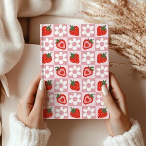 Hands holding a strawberry and daisy patterned hardcover notebook with a pink and red checkered background. The person is wrapped in a cream sweater.