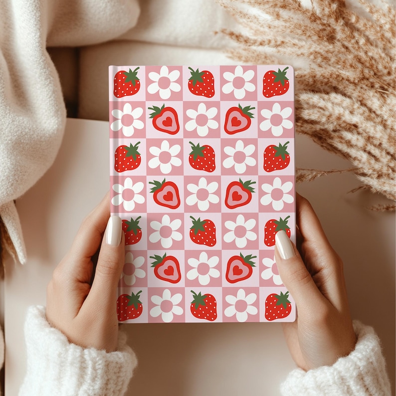 Hands holding a strawberry and daisy patterned hardcover notebook with a pink and red checkered background. The person is wrapped in a cream sweater.