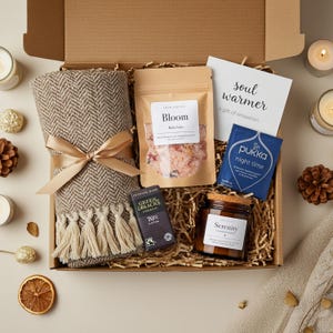 May include: A gift box containing a beige herringbone blanket, bath salts, a dark chocolate bar, a candle, and a tea bag. The box also includes a card that reads "soul warmer, a gift of relaxation". The candle is labelled "Serenity".