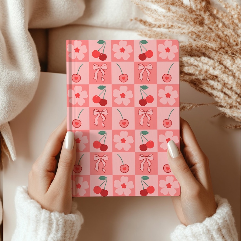 Hands holding a cherry-themed hardcover notebook with alternating squares of bows, hearts, and flowers in a soft pink color palette.