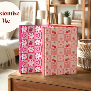 Two custom hardcover notebooks standing on a wooden table in a cozy room. One has a raspberry, heart, and flower pattern with the spine text "Geography Notes", and the other features cherries, bows, and flowers with the text "Alice's Notes". A circular label reads “Customise Me”.