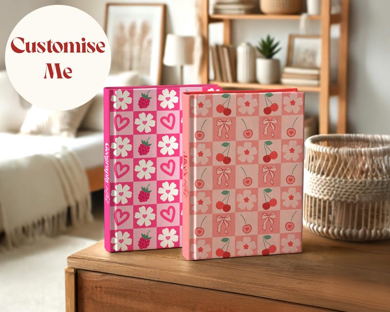 Two custom hardcover notebooks standing on a wooden table in a cozy room. One has a raspberry, heart, and flower pattern with the spine text "Geography Notes", and the other features cherries, bows, and flowers with the text "Alice's Notes". A circular label reads “Customise Me”.